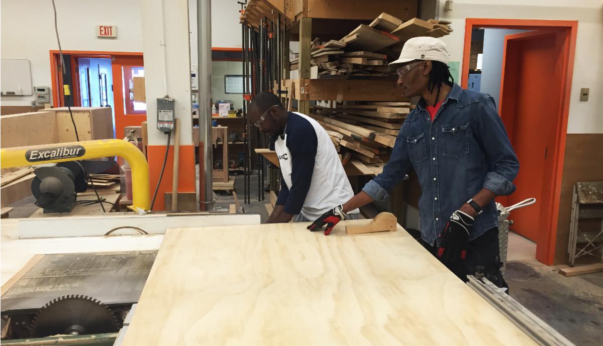 Two people stand in a full workshop, in front of a large piece of plywood.