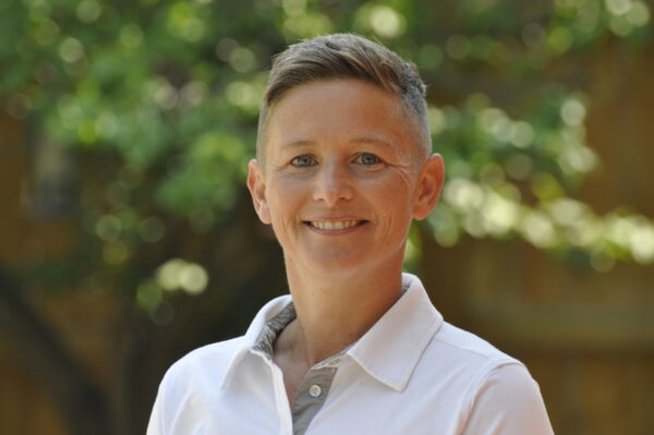 A headshot of Sarah Aspinall. She has short hair and wears a white collared shirt. She is standing outside in front of greenery.