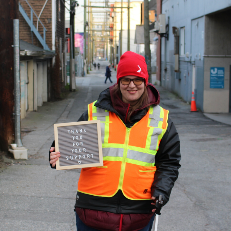 A person wearing a visi-vest and red toque holds a sign that says Thank you for your support. They stand in an alley.