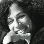 Black and white portrait of Nancy Neamtan. She has curly hair, and rests her chin on her hand as she smiles.